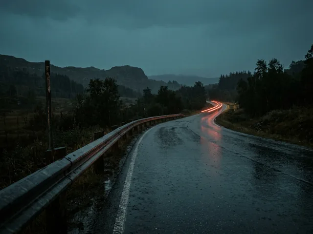An empty wet road at dusk with one red tail light streaking away into the rain.