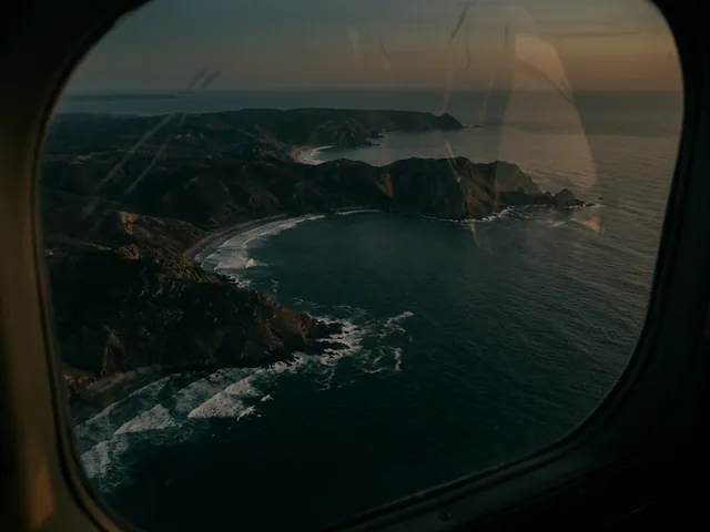 A coastline seen from an aircraft window at dusk, headlands and pale surf lines below.