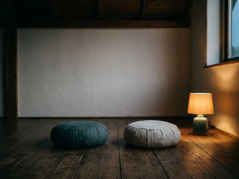 Two round meditation cushions on a bare wooden floor beside a low lamp