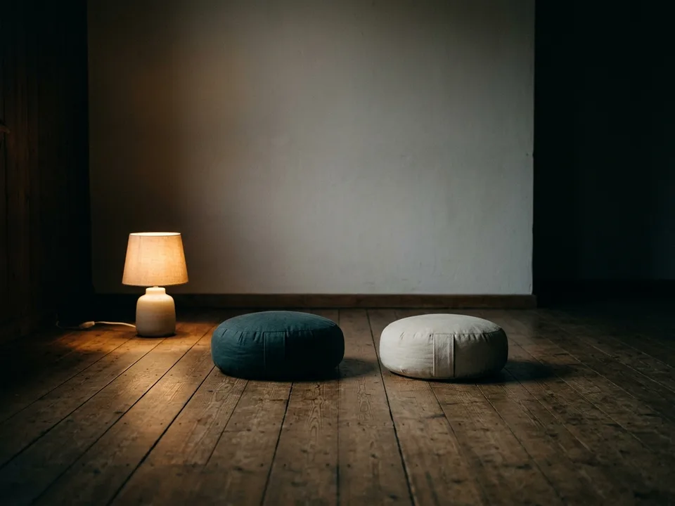 Two round meditation cushions on a bare wooden floor beside a low lamp
