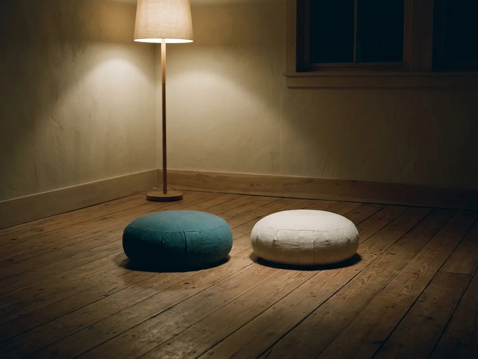 Two round meditation cushions on a bare wooden floor beside a low lamp