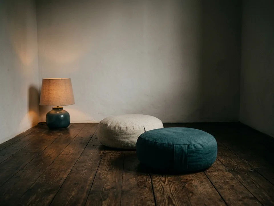 Two round meditation cushions on a bare wooden floor beside a low lamp