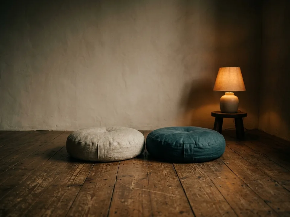 Two round meditation cushions on a bare wooden floor beside a low lamp
