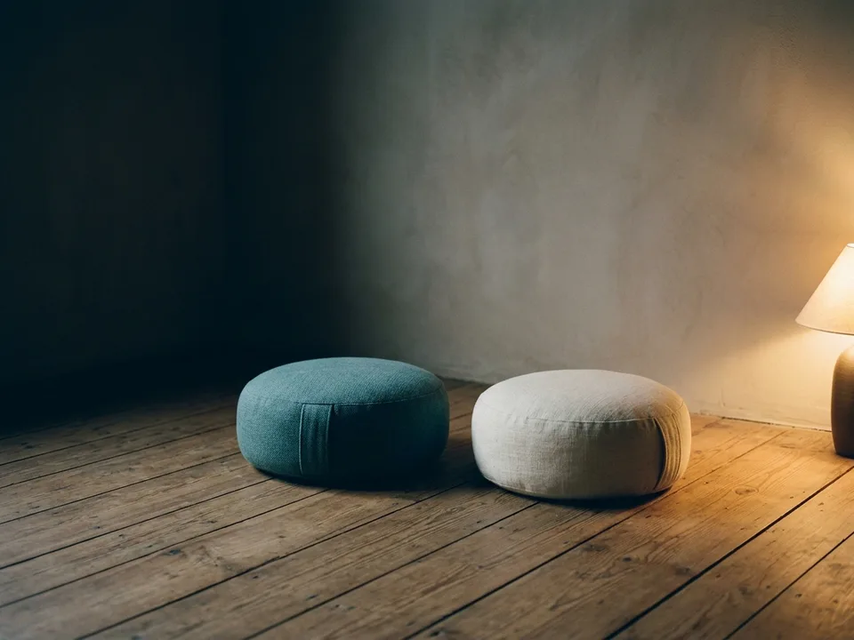 Two round meditation cushions on a bare wooden floor beside a low lamp